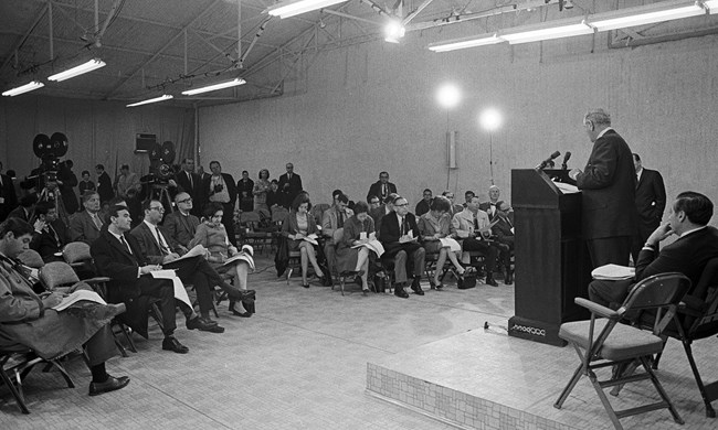 Black and white photo of President Johnson speaking to a crowd of reporters and photographers.