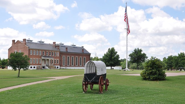 A large brick two-story building sits in the background.  A covered wagon and flagpole flying an American flag are seen in the forefront of the photo.