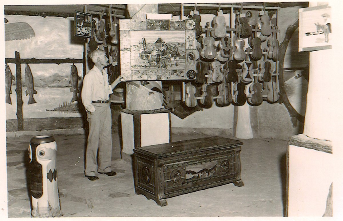 Fiddle House Man standing open room next to picture and ornate trunk at his feet. Over 50 fiddles hang on wall in background.