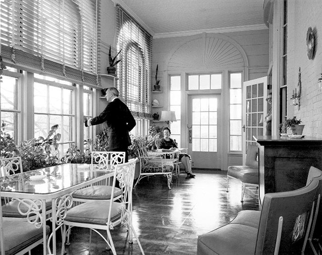 Interior Porch of the Commandant's House with a man looking out the window and a woman relaxing in a chair.