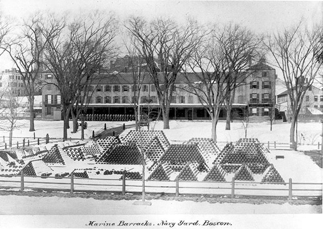 Piles of cannon balls in the foreground covered in snow with a snowy parade ground and Marine Barracks in the background.