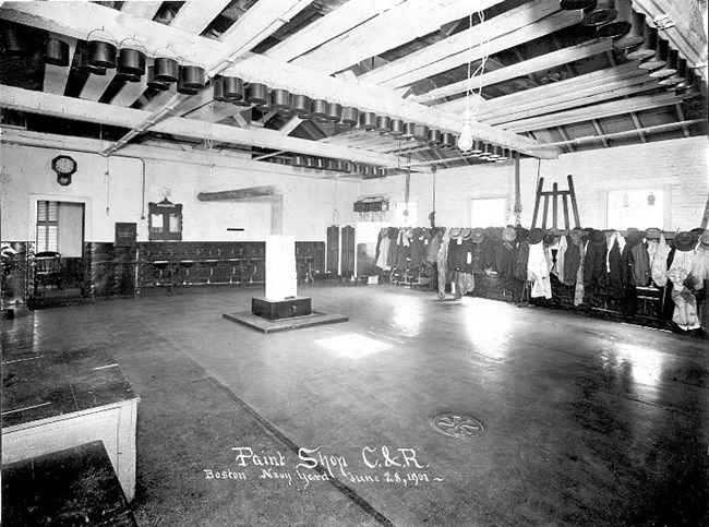 A large open room with jackets hanging up along a wall. Caption identifies it as the paint shop.