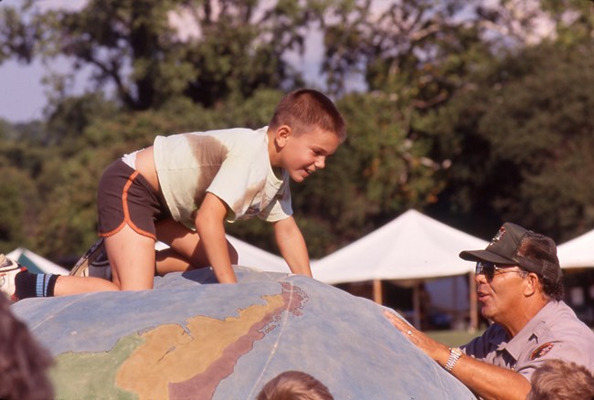Child climbs atop a large inflated globe supported by a man in a ranger uniform. Behind them are event tents.