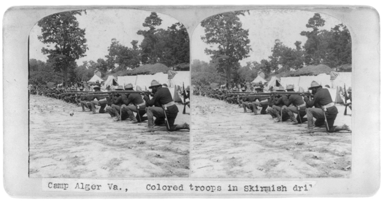 Black and white photo of African American soldiers kneeling in skirmish drill with rifles to their shoulders.