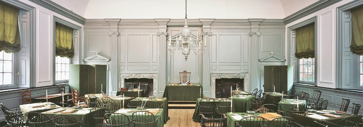 Thirteen tables with green tablecloths arranged in a semi-circle with an ornate glass chandilier hanging from the ceiling.