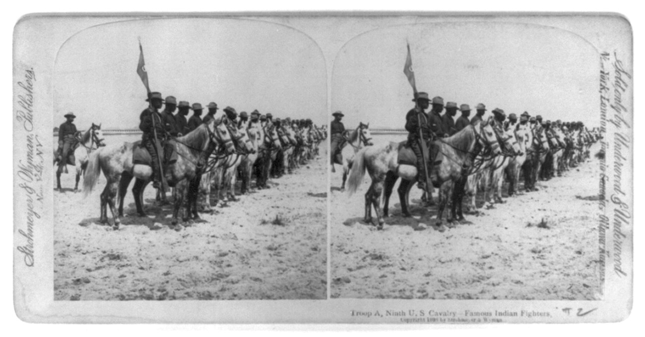 Stereograph photo of African American Soldiers lines up shoulder to shoulder mounted on their horses. The Soldier nearest the viewer is holding the unit flag on a pole.