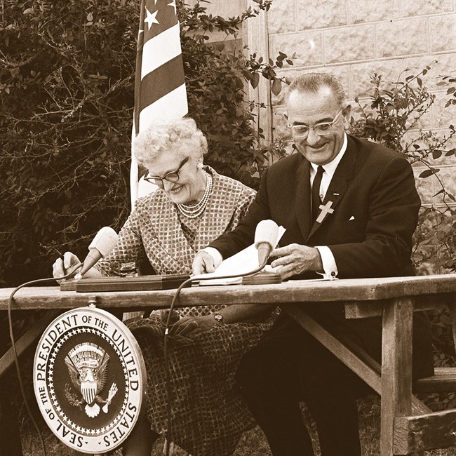 A smiling elderly lady in checkered dress sits next to a smiling gentleman in a suit. There is a presidential seal in front of the picnic table where they sit.