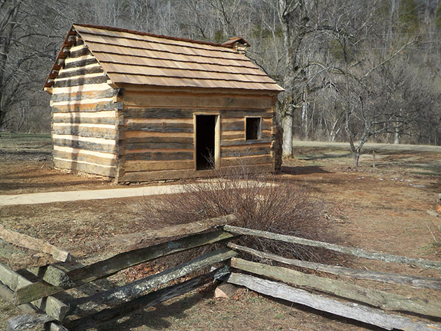 A small rustic log cabin with a wooden shingle roof stands in a clearing surrounded by leafless trees