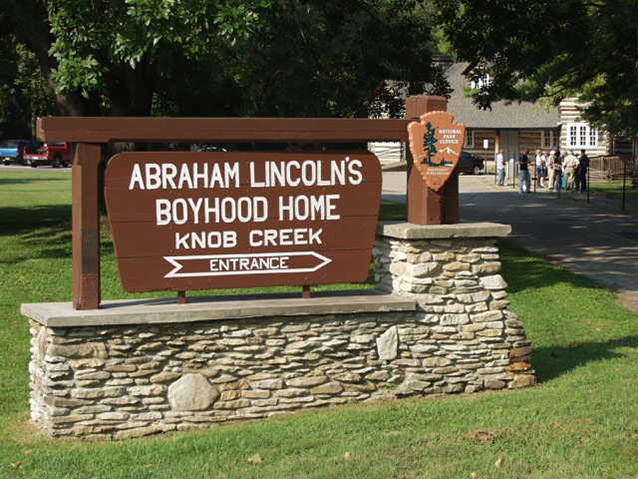 A large wooden sign on a stone base reads “Abraham Lincoln’s Boyhood Home, Knob Creek, Entrance,” with an arrow pointing right.