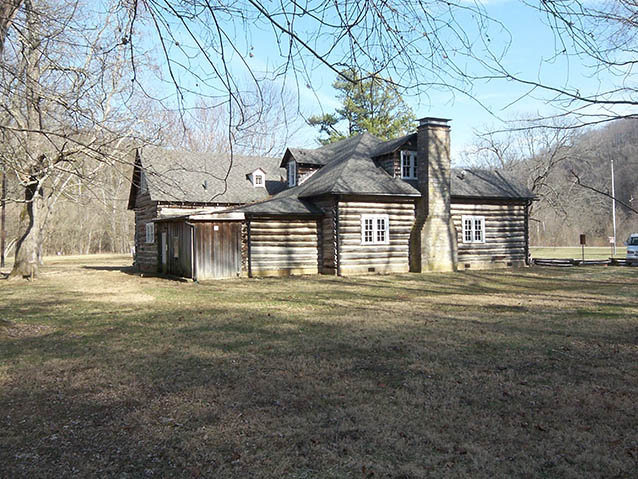 A large historic log building with multiple rooflines and a stone chimney sits in an open grassy area surrounded by leafless trees on a clear day.