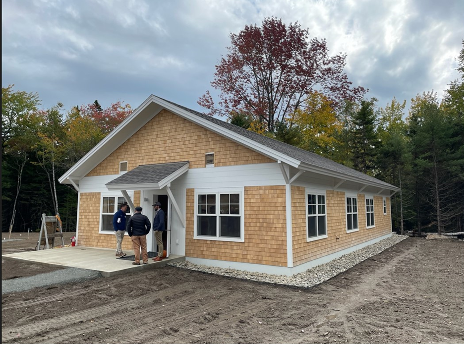 Dane Farm Housing at Acadia National Park Prototype house with three people standing around front door with gravel in front and deciduous tress visible in the background