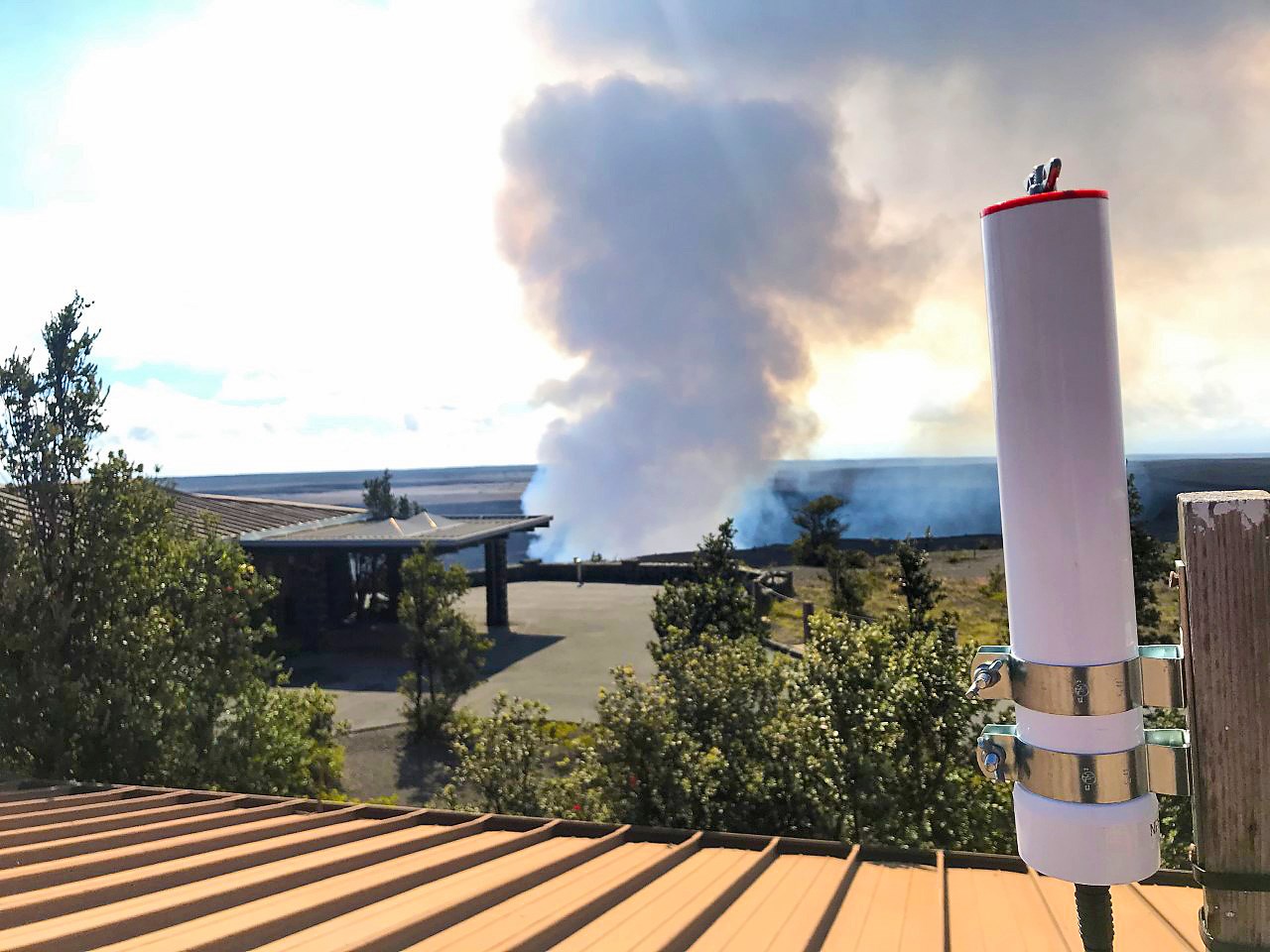 Flight track recording unit White cylyndrical device mounted to a post on a rooftop overlooking Kīlauea Volcano as it spews a plume of gasses into the air.