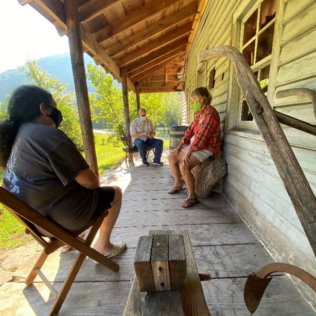 Three people seated on wooden porch of historic wood home with heritage outdoor furniture