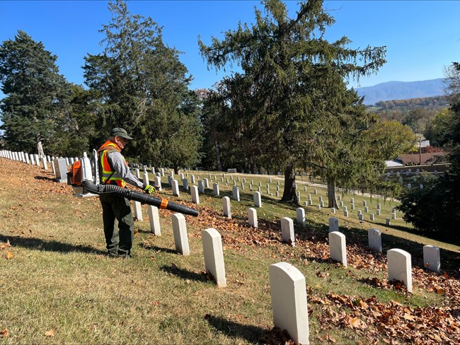 AJ uses a leaf blower to remove leaves in section E of Cemetery Andrew Johnson National Historic Site. Maintenance worker stands near headstones in a cemetary on a sunny day using a leaf blower to remove leaves from the area.