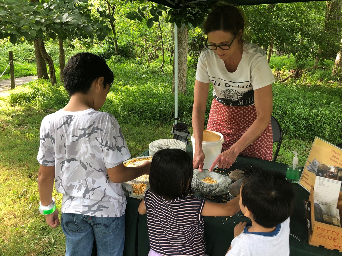 A woman shows three children how corn is ground using a mortar and pestle