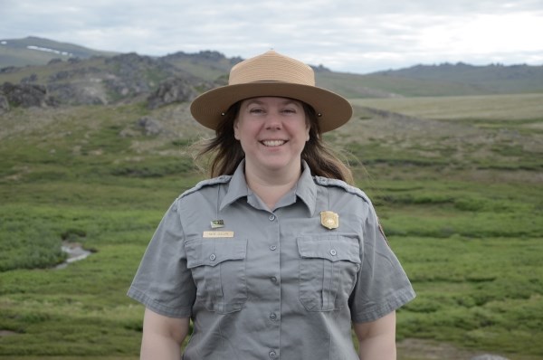 Katie poses for a picture wearing a national park service uniform, in the background mountains are in the distance.