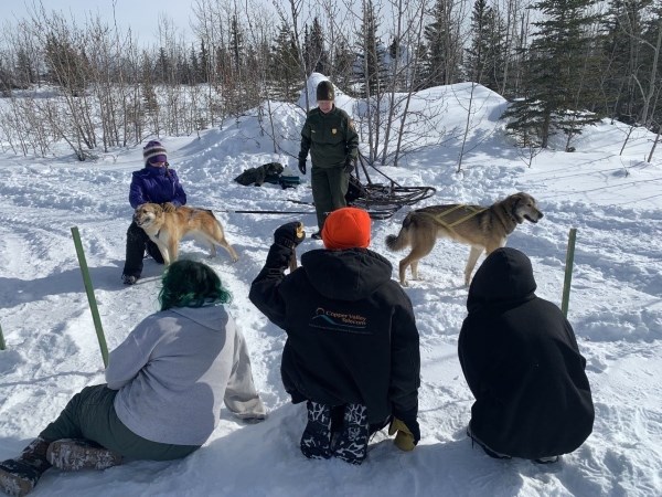 Three children sit in the snowy landscape as a park ranger in uniform stands by a dog sled with two sled dogs in harness. A person to the left of the ranger holds on to a dog in harness.