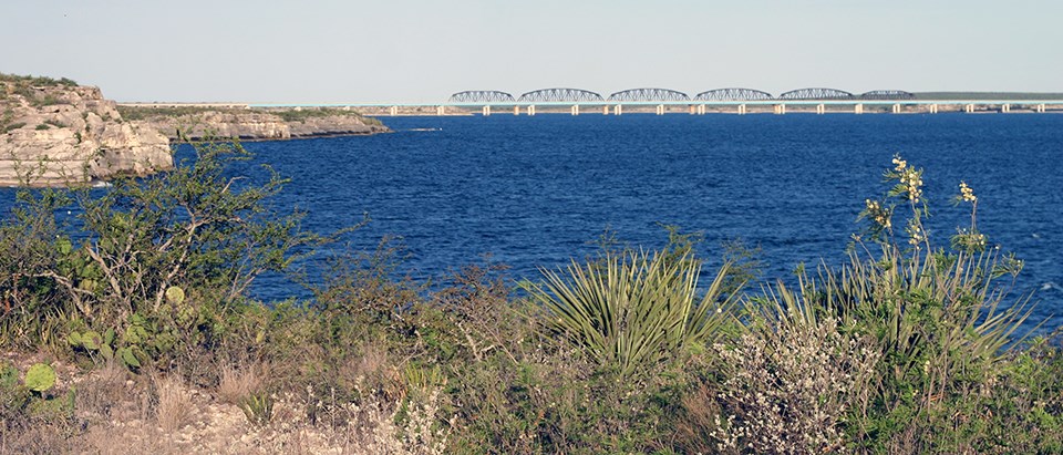 A large, rippling, blue lake lined by sheer rock cliffs topped by desert vegetation and a long bridge in the distance.