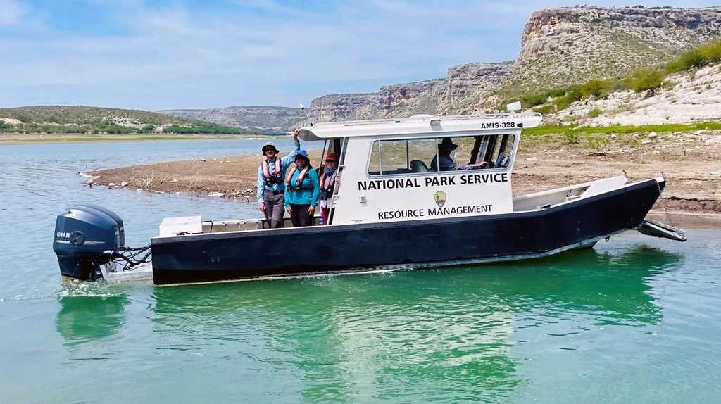 Amistad Reservoir at Amistad National Recreation Area Four park staff members on a boat next to the shore of a reservoir surrounded by desert hills. The boat has writing that says, "National Park Service Resource Management."