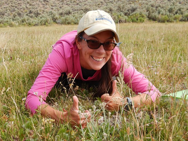 Jackie Lucero Smiling woman crouched in a field giving the thumbs up.