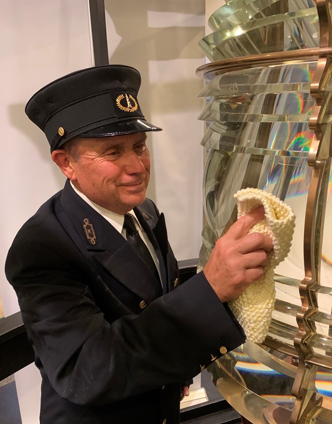 Light keeper polishing the Fresnel Lens. A man dressed in a black lighthouse keeper uniform and hat polishes a glass Fresnel lens.
