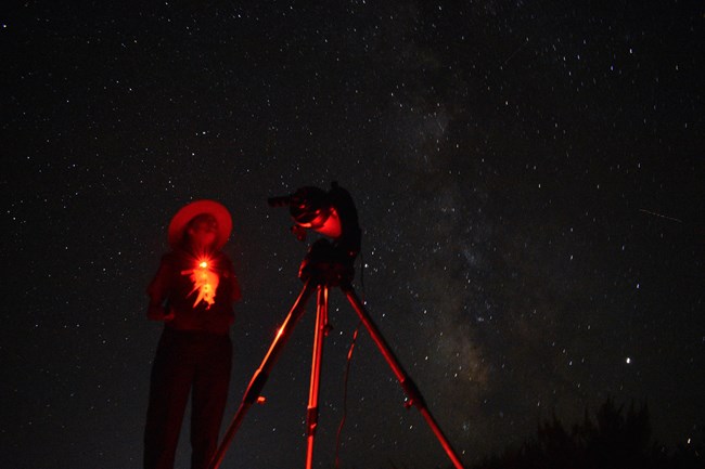 a ranger and a telescope illuminated in red with a starry sky overhead