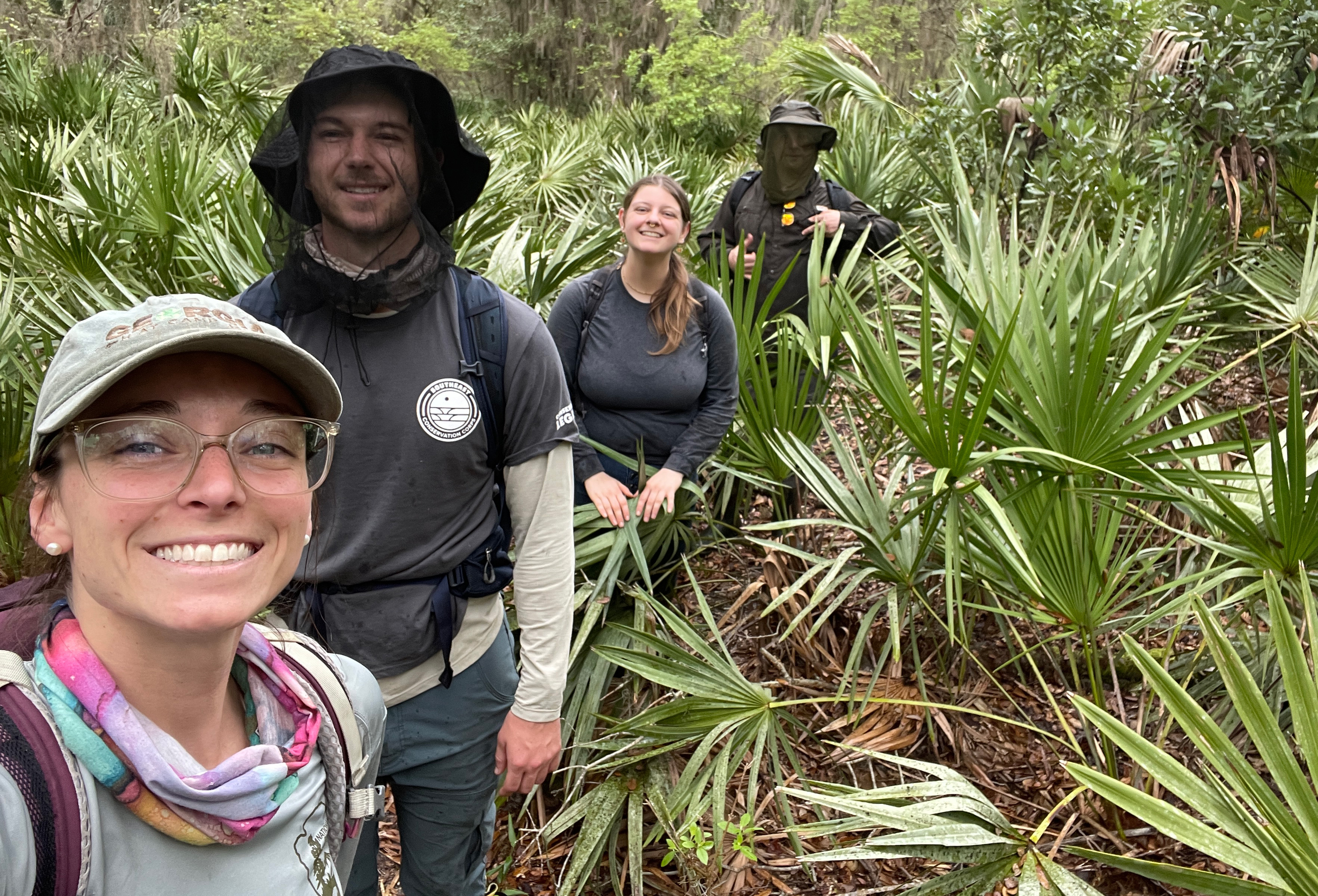 People standing in bushes and palmettos smiling. One wears a mosquito net.