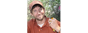 Ian in a brick red shirt and ball cap, holding a black and bright orange bird in one hand.