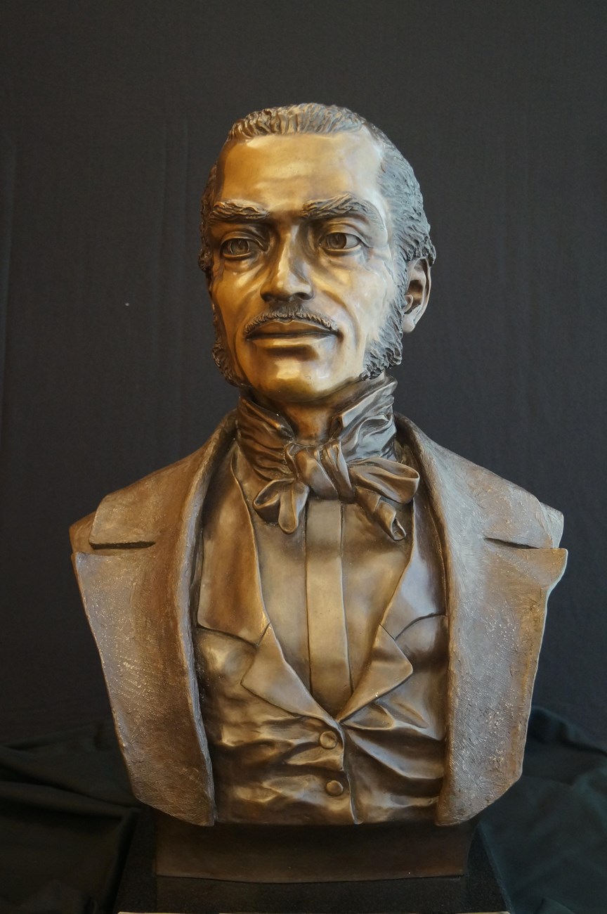 Bust of a Black man named Free Frank McWorter. This bust appears to be made of copper or bronze. Frank looks slightly to his right with a stoic expression on his face. He wears a suit jacket, a collared shirt, a vest, and a cravat.