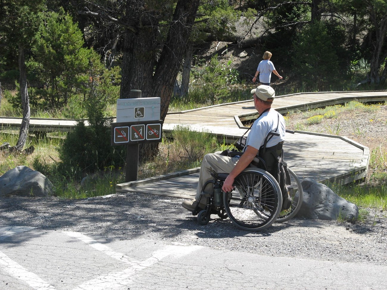 Man in manual wheelchair in front of a wooden trail sign featuring the international symbol of access in front of a wooden boardwalk. A woman walks along the boardwalk in the background of the photo.