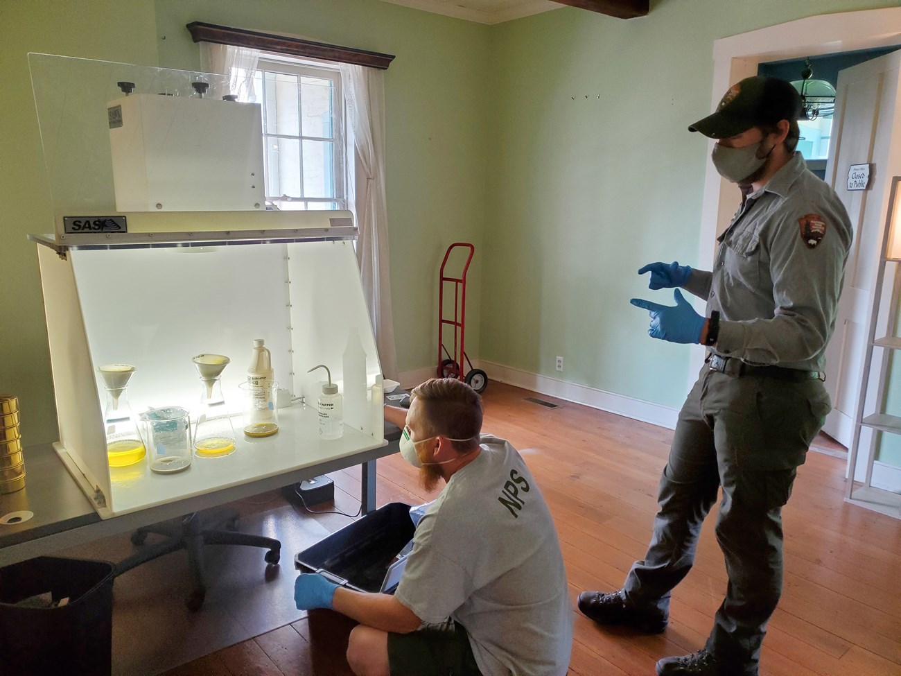 Watching over acid digestion at Sainte Genevieve National Historical Park Two men with masks and NPS uniforms look at chemicals in glassware stored inside a fume hood.