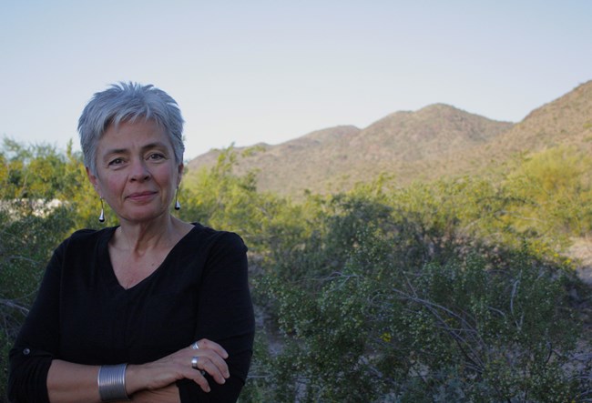 Woman with short light hair and silver earrings, rings, and a bracelet stands outdoors with mountains in distance