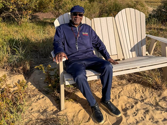 An African American man with blue pants, jacket, and hat sits on a bench.