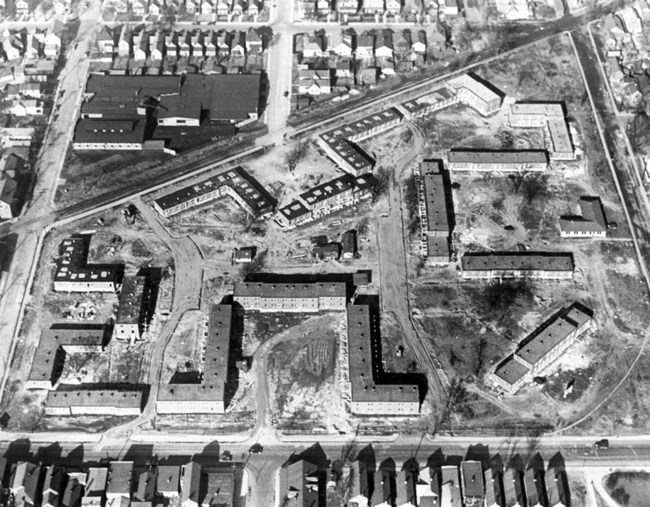 Sixteen L-shaped and long rectangular buildings on large, irregular-shaped construction site. Dirt paths and ground cover are still visible. The development is across from a large high school building and surrounded by housing.