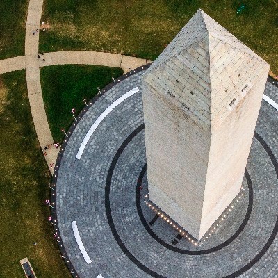 Aerial view looking down on a tall white stone obelisk in the center of a large circle of gray brick in a grassy park.