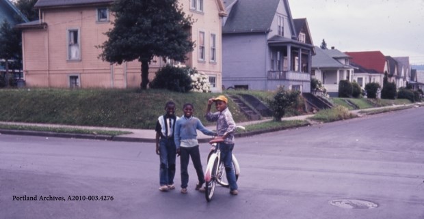 3 African American boys stand around a bicycle in a neighborhood road.
