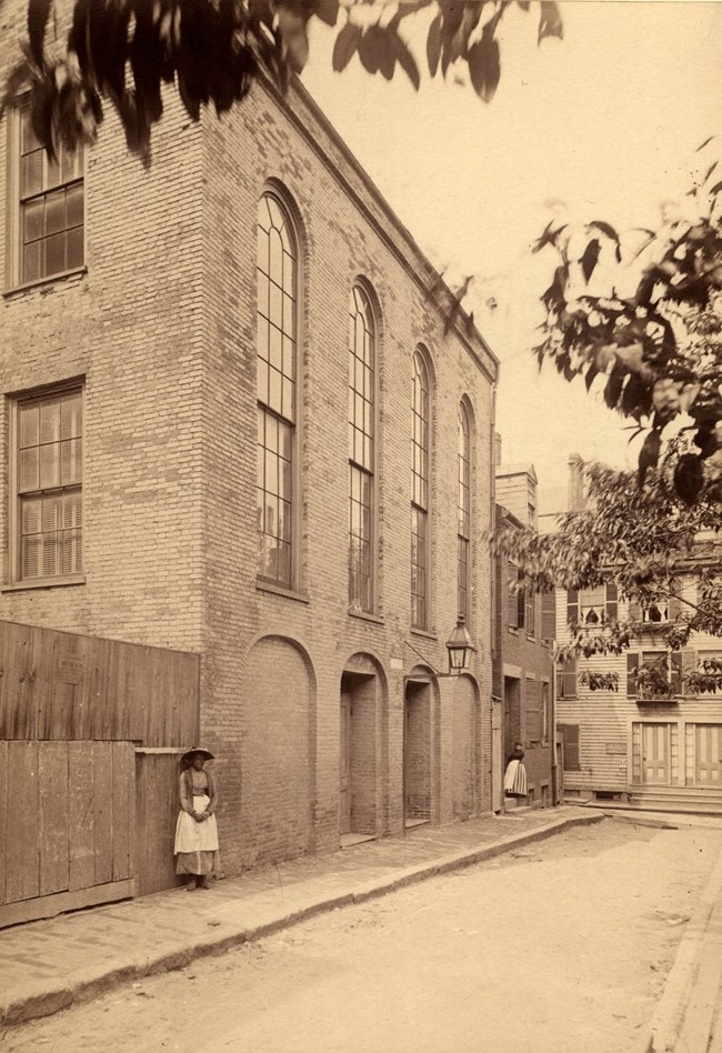 AfricanMeetingHouse-MHS Woman stands in front of brick three-story building with tall arched windows.