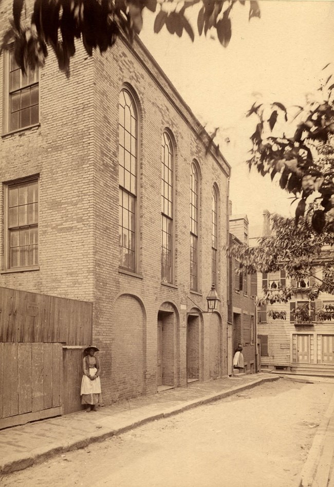 Woman stands in front of brick three-story building with tall arched windows.