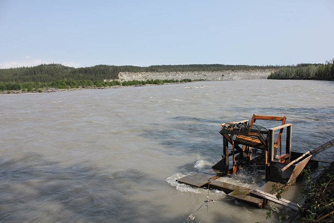 Ahtna.Fishwheel on River_web A wide river with a fishwheel along one bank.