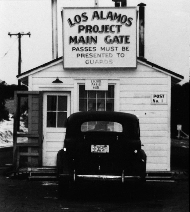 Black and white photo of the back of a dark car parked at a white clapboard building under a sign "Los Alamos Project Main Gate"