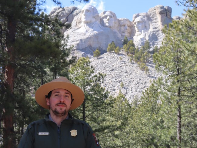 A smiling man with a brown goatee wears a NPS flat hat, badge, and nametag while standing in front of the sculpture known as Mount Rushmore.