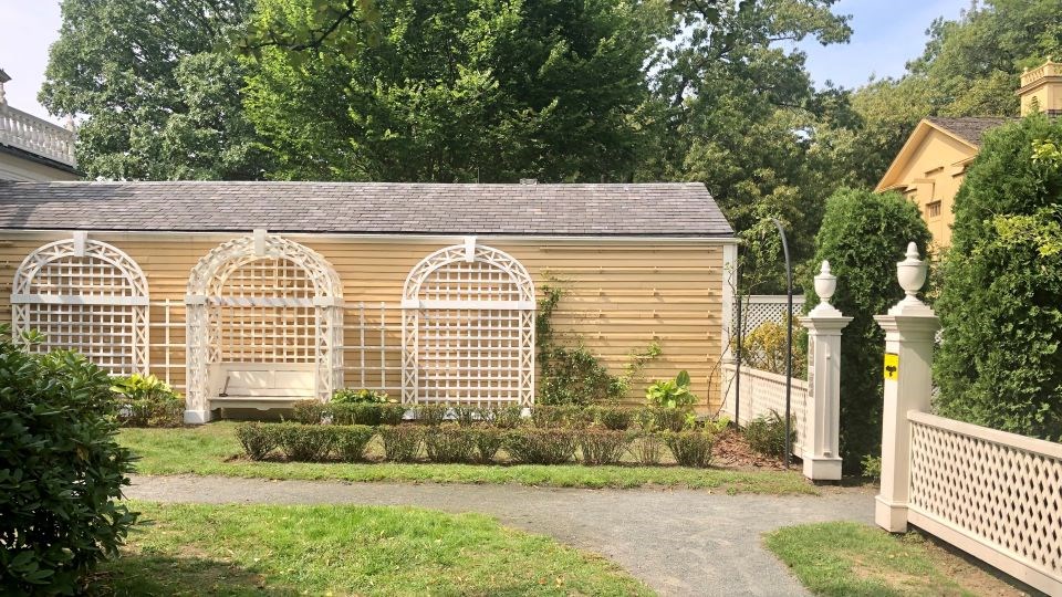 Garden area featuring three white arched trellises against yellow clapboard wall with low bushes in front.