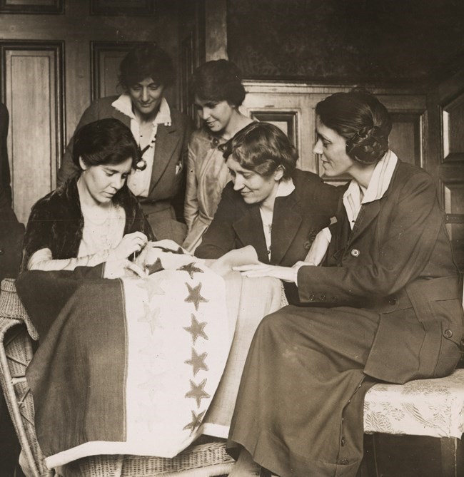 A woman sewing a flag. Four other women watch her.