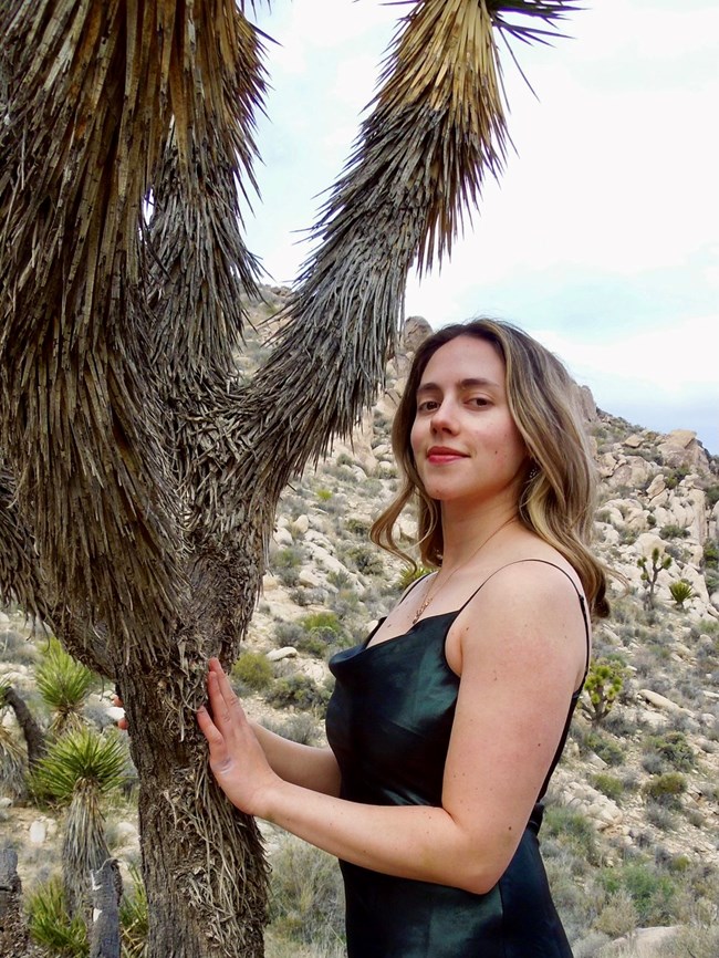 A woman in a black tanktop holds her hands against the trunk of a tree while looking at us.