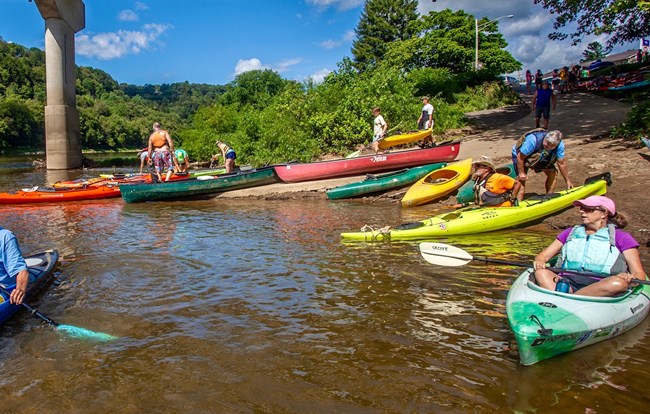 Kayakers and canoers head out onto river from boat launch. Blue, sunny skies and lush green trees on riverbank.
