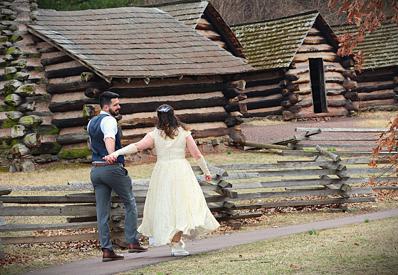 A bride and a groom hold hands as they walk towards wooden huts.