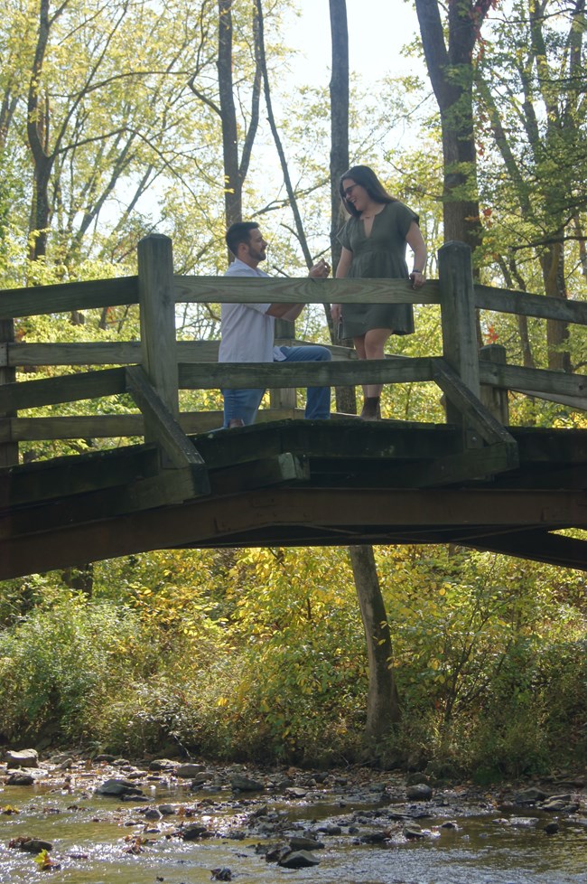 A person on one knee proposing to another on a wooden bridge over a creek.