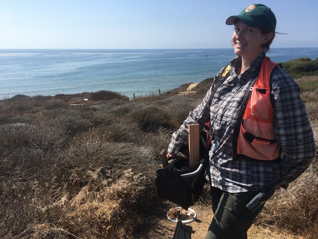 Ambassador Samantha Wynns 2 Woman in field clothes, orange safety vest, and green ballcap with the NPS arrowhead on it stands smiling in front of the green guide fencing of a herpetofauna pitfall array at Cabrillo National Monument. Blue ocean stretches to the horizon behind her.