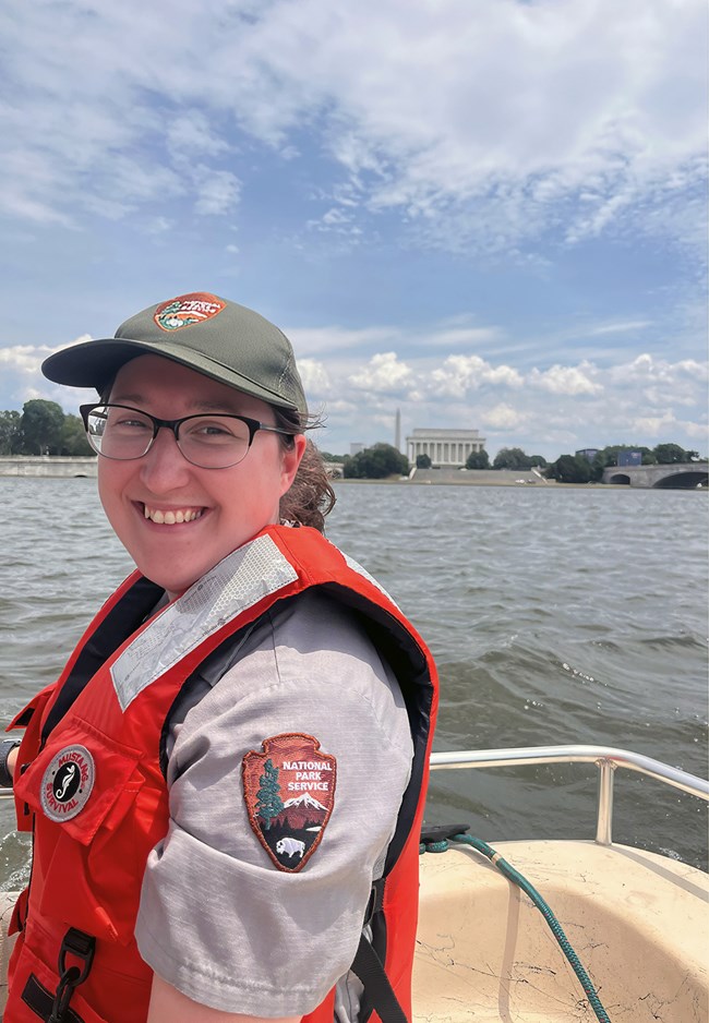 A person in a ball cap and glasses, sits in a boat on a flat river.
