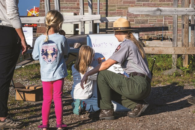 A park ranger kneels on the ground while a child writes on a whiteboard next to them.